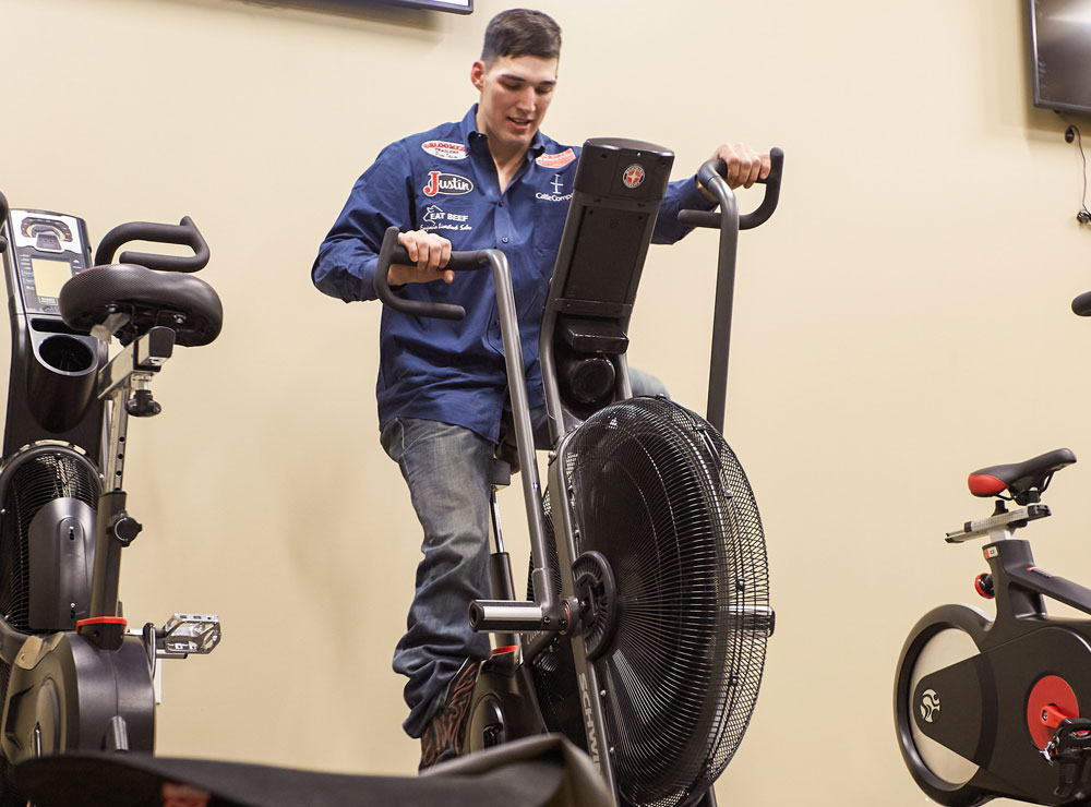 Jess Pope is wearing a blue button-down shirt and blue jeans, riding a stationary bike to warm up before competing at a rodeo. 